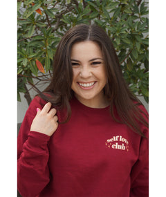A woman modeling a red crewneck sweatshirt with “Self love” in white serif font outlined in orange arched above “club” in white serif font outline in orange. “Club” is surrounded by orange stars - from Sierra Schultzzie