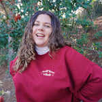 A woman modeling a red crewneck sweatshirt with “Self love” in white serif font outlined in orange arched above “club” in white serif font outline in orange. “Club” is surrounded by orange stars - from Sierra Schultzzie