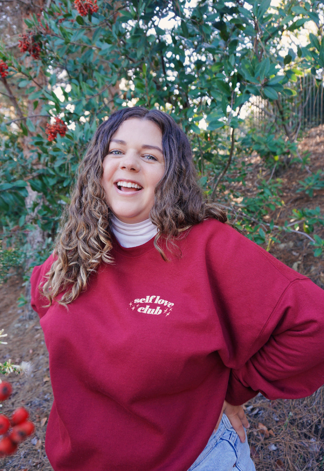 A woman modeling a red crewneck sweatshirt with “Self love” in white serif font outlined in orange arched above “club” in white serif font outline in orange. “Club” is surrounded by orange stars - from Sierra Schultzzie