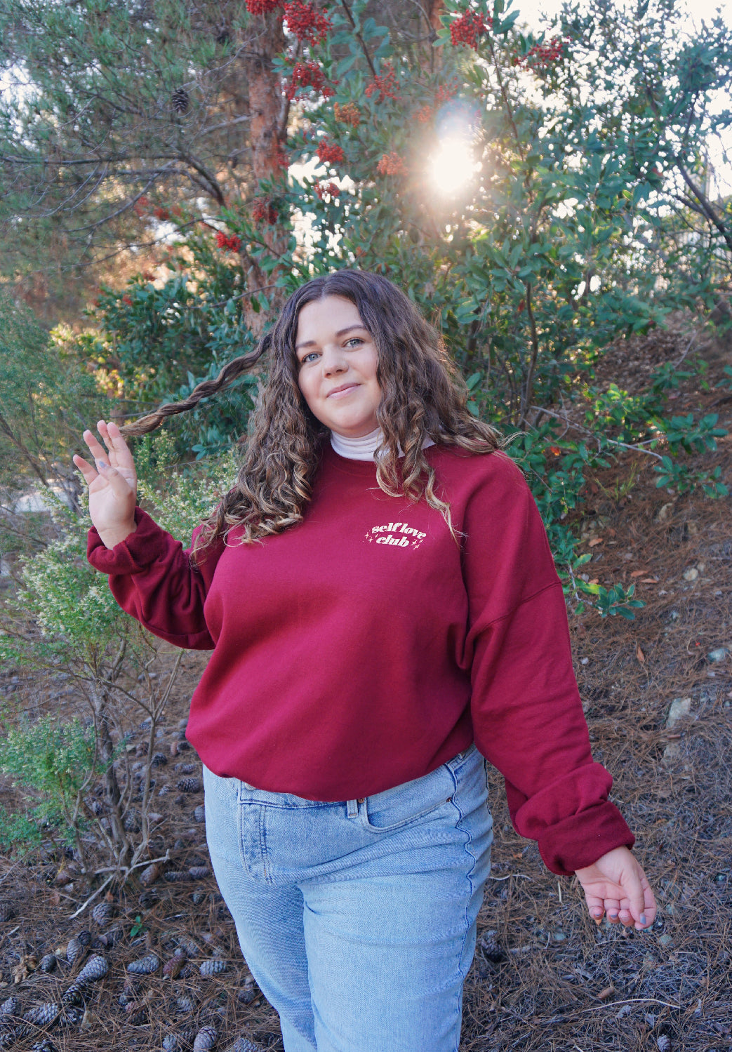 A woman modeling a red crewneck sweatshirt with “Self love” in white serif font outlined in orange arched above “club” in white serif font outline in orange. “Club” is surrounded by orange stars - from Sierra Schultzzie
