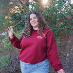 A woman modeling a red crewneck sweatshirt with “Self love” in white serif font outlined in orange arched above “club” in white serif font outline in orange. “Club” is surrounded by orange stars - from Sierra Schultzzie