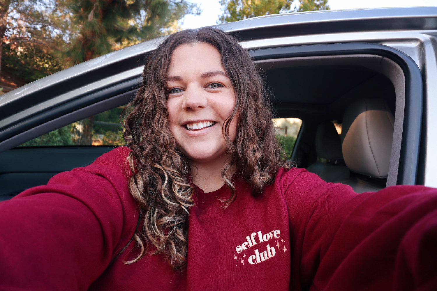 A woman modeling a red crewneck sweatshirt with “Self love” in white serif font outlined in orange arched above “club” in white serif font outline in orange. “Club” is surrounded by orange stars - from Sierra Schultzzie