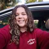 A woman modeling a red crewneck sweatshirt with “Self love” in white serif font outlined in orange arched above “club” in white serif font outline in orange. “Club” is surrounded by orange stars - from Sierra Schultzzie