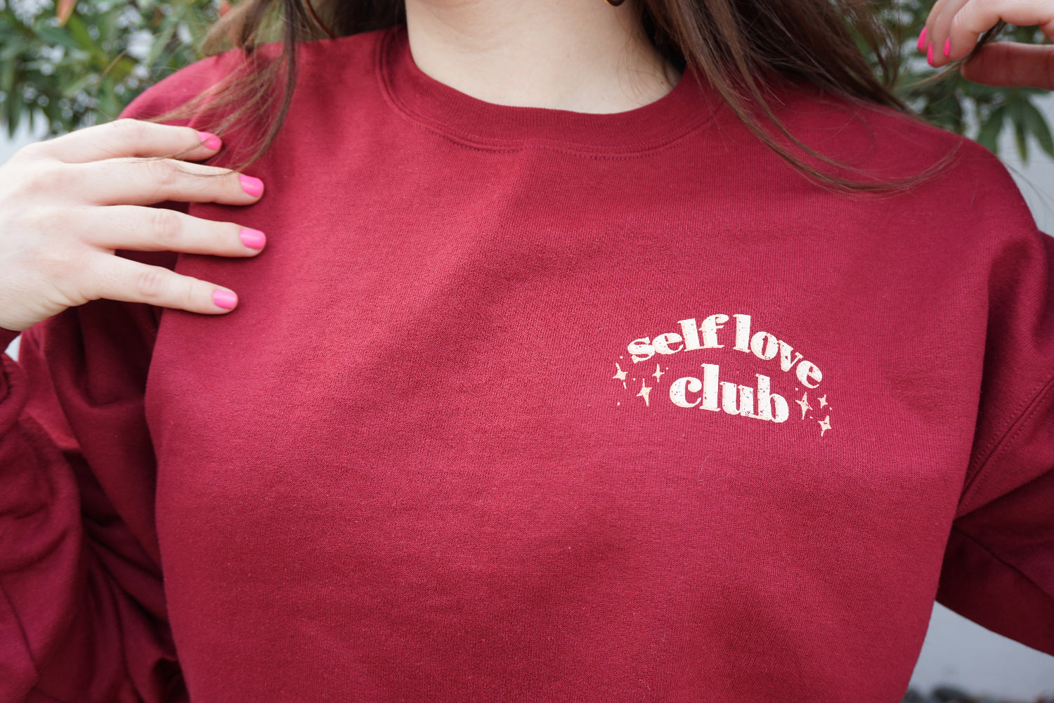 A woman modeling a red crewneck sweatshirt with “Self love” in white serif font outlined in orange arched above “club” in white serif font outline in orange. “Club” is surrounded by orange stars - from Sierra Schultzzie