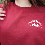 A woman modeling a red crewneck sweatshirt with “Self love” in white serif font outlined in orange arched above “club” in white serif font outline in orange. “Club” is surrounded by orange stars - from Sierra Schultzzie