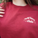 A woman modeling a red crewneck sweatshirt with “Self love” in white serif font outlined in orange arched above “club” in white serif font outline in orange. “Club” is surrounded by orange stars - from Sierra Schultzzie