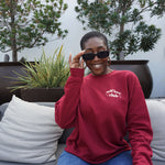 A woman modeling a red crewneck sweatshirt with “Self love” in white serif font outlined in orange arched above “club” in white serif font outline in orange. “Club” is surrounded by orange stars - from Sierra Schultzzie