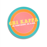 A circular sticker with a colorful, ringed background that says "Chi Kappa; Be confident. Be kind." From Sierra Schultzzie.