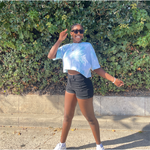 Someone wearing the blue confident and kind crop top from Sierra Schultzzie, jean shorts and sunglasses, and facing the camera smiling.