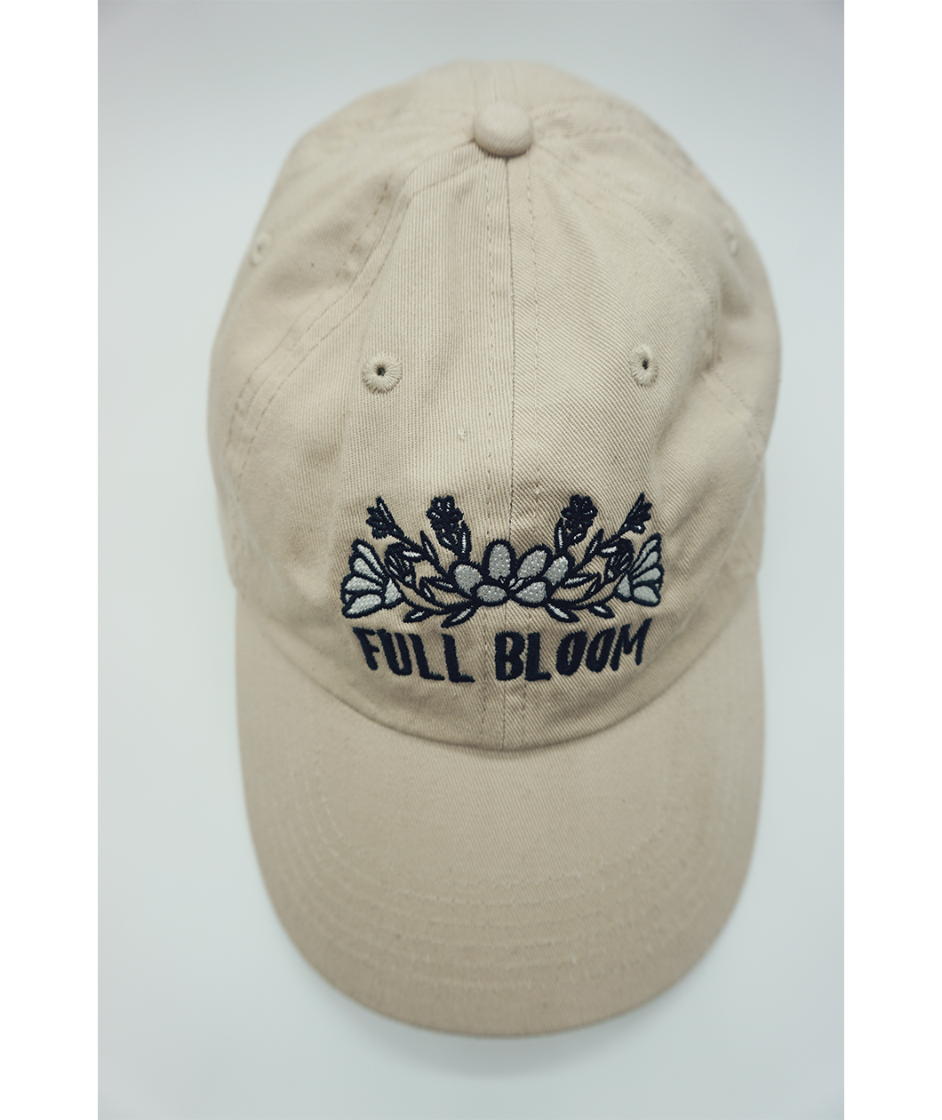 A top view of a beige ball cap with the embroidered words "Full Bloom" and flowers from Sierra Schultzzie.