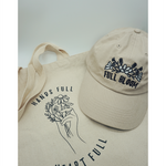 Close up of the canvas tote from Sierra Schultzzie with the words "Hands Full, Heart Full" printed in black as well as an outline of a hand holding flowers. On top of the tote in a beige ball cap with the embroidered words "Full Bloom" and flowers.