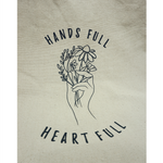 Close up of the canvas tote from Sierra Schultzzie with the words "Hands Full, Heart Full" printed in black as well as an outline of a hand holding flowers.