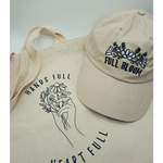 Close up of the canvas tote from Sierra Schultzzie with the words "Hands Full, Heart Full" printed in black as well as an outline of a hand holding flowers. On top of the tote in a beige ball cap with the embroidered words "Full Bloom" and flowers.