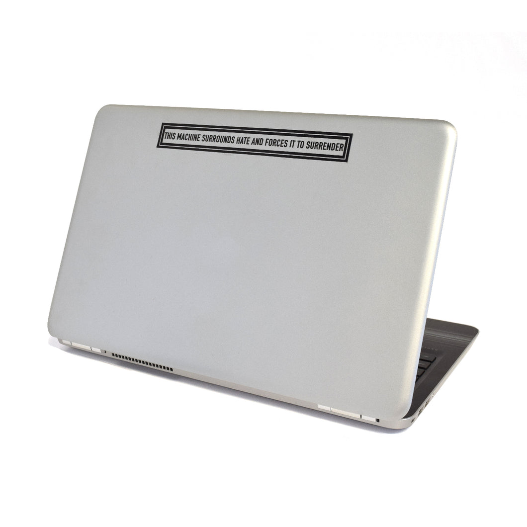 A gray laptop with a long rectangular sticker with two black borders surrounding “This Machine Surrounds Hate and Forces It To Surrender” in black sans serif font at the top of the laptop - from John Green