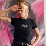 A photo of Catherine McCafferty posing, wearing a black t-shirt. The shirt reads "Pretty Gay" and has little star illustrations around it. From Pretty Gay.