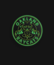 Green and black circular pin centering a cat with the phrase "Oakland Raycats" around it - by 99% Invisible. Parts of it glow in in dark in bright green.