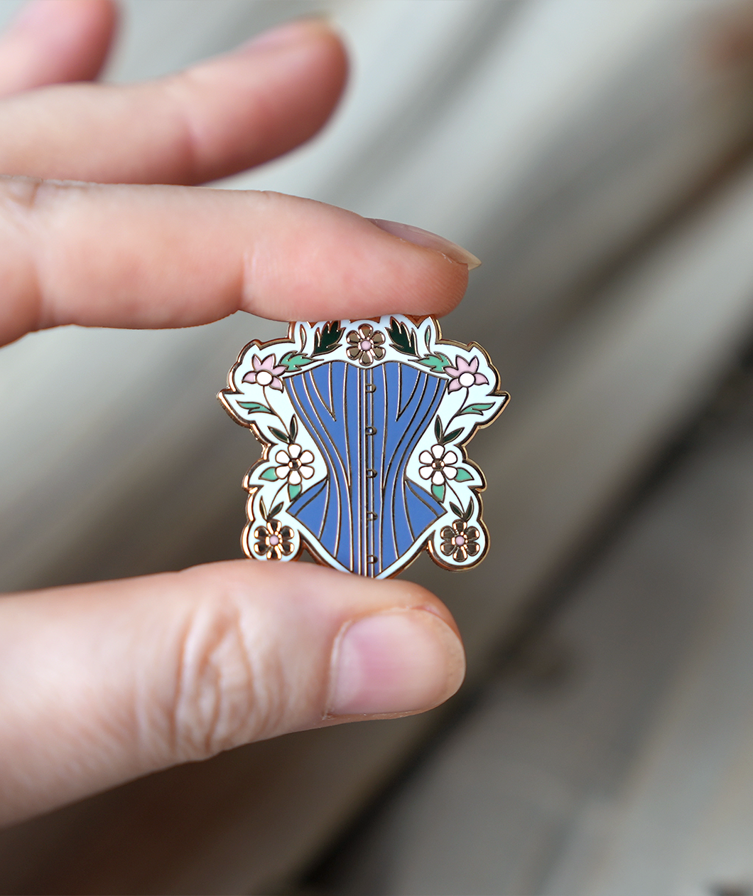 Gold enamel pin held between someone's thumb and forefinger to show size. Pin is about 1.25" tall and depicts a powder blue corset surrounded by gold, white, and pink daises with green leaves and a light blue background.