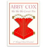 Gold hard enamel pin shaped like a corset filled with red enamel and a white enamel top edge to look like lace with gold detailing. The pin is split down the middle with golden hinges on each of the four corners to show it can open. The pin is placed on a cream colored backing card that says ABBY COX in pink lettering on top. Right below it says Ho Ho Ho Corset Pin. Under the pin the backer card says 2" Hinged Hard Enamel Pin.