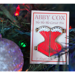 Gold hard enamel pin shaped like a corset filled with red enamel and a white enamel top edge to look like lace with gold detailing. The pin is split down the middle with golden hinges on each of the four corners to show it can open. Packaged on a cream colored backer card that says, "ABBY COX Ho Ho Ho Corset Pin 2" Hinged Hard Enamel". Seen in a Christmas tree next to a sparkly ornament.