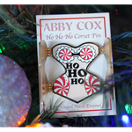 Gold hard enamel pin has either side opened to reveal the words "HO HO HO" in the center with candy cane patterned padding on each hip and a peppermint bra with white enamel lace. Small magnets are on the back of the two open front flaps. Packaged on a cream colored backer card that says, "ABBY COX Ho Ho Ho Corset Pin 2" Hinged Hard Enamel". Seen in a Christmas tree next to a sparkly ornament.