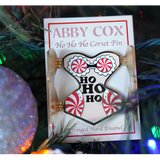 Gold hard enamel pin has either side opened to reveal the words "HO HO HO" in the center with candy cane patterned padding on each hip and a peppermint bra with white enamel lace. Small magnets are on the back of the two open front flaps. Packaged on a cream colored backer card that says, "ABBY COX Ho Ho Ho Corset Pin 2" Hinged Hard Enamel". Seen in a Christmas tree next to a sparkly ornament.