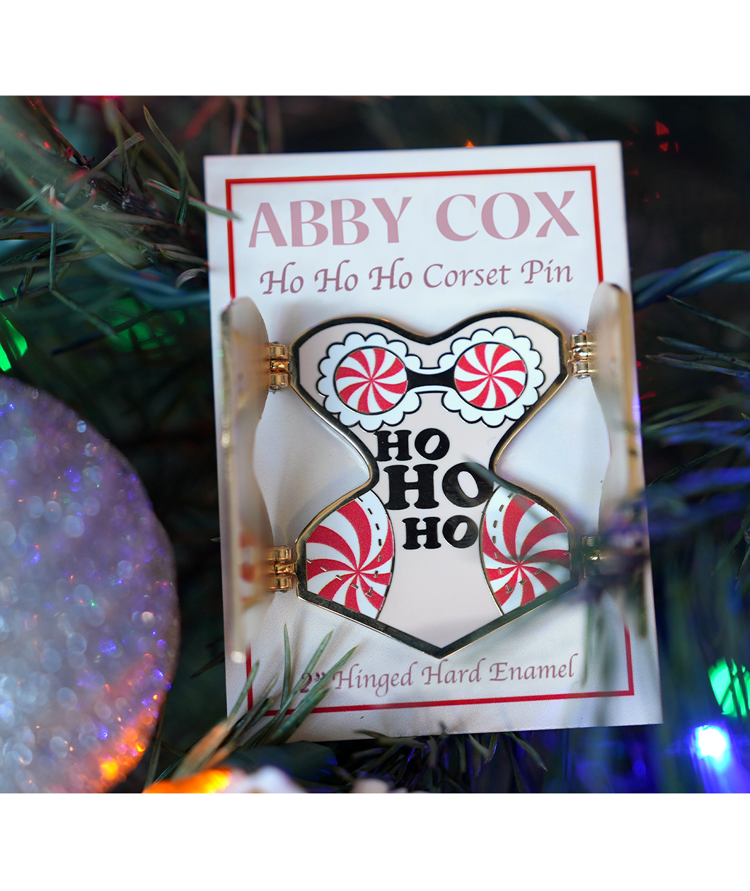 Gold hard enamel pin has either side opened to reveal the words "HO HO HO" in the center with candy cane patterned padding on each hip and a peppermint bra with white enamel lace. Small magnets are on the back of the two open front flaps. Packaged on a cream colored backer card that says, "ABBY COX Ho Ho Ho Corset Pin 2" Hinged Hard Enamel". Seen in a Christmas tree next to a sparkly ornament.