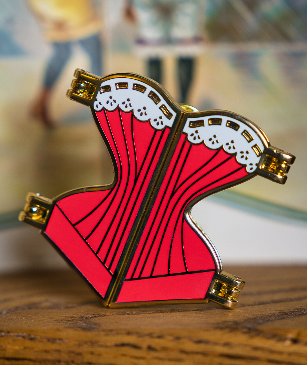 Gold hard enamel pin shaped like a corset filled with red enamel and a white enamel top edge to look like lace with gold detailing. The pin is split down the middle with golden hinges on each of the four corners to show it can open.