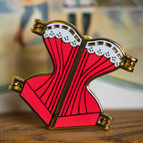 Gold hard enamel pin shaped like a corset filled with red enamel and a white enamel top edge to look like lace with gold detailing. The pin is split down the middle with golden hinges on each of the four corners to show it can open.