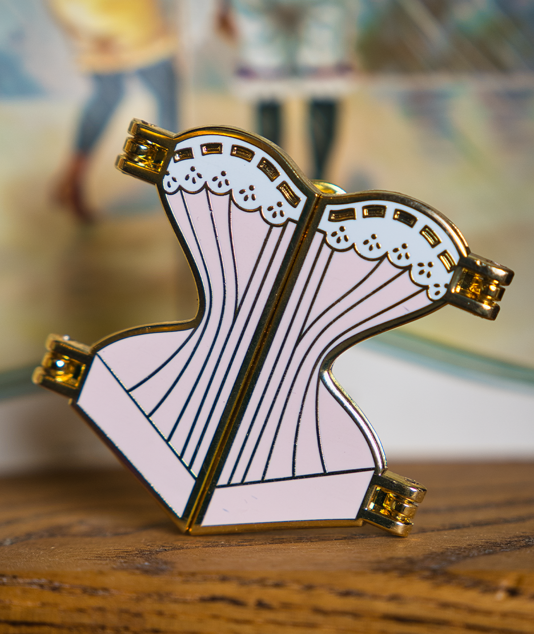 Gold hard enamel pin shaped like a corset filled with pale pink enamel and a white enamel top edge to look like lace with gold detailing. The pin is split down the middle with golden hinges on each of the four corners to show it can open.