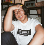 A photo of person sitting on the ground, smiling with their eyes closed wearing a white t-shirt with a black rectangle in the center with the words "We're figuring it out" written three times in a row with the second line has "figuring" crossed out and the third line has "we're" and "it" crossed out. There is additional small text below this. From Answer in Progress.