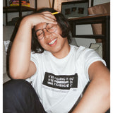 A photo of person sitting on the ground, smiling with their eyes closed wearing a white t-shirt with a black rectangle in the center with the words "We're figuring it out" written three times in a row with the second line has "figuring" crossed out and the third line has "we're" and "it" crossed out. There is additional small text below this. From Answer in Progress.