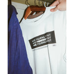A photo of person hanging up a white t-shirt with a black rectangle in the center with the words "We're figuring it out" written three times in a row with the second line has "figuring" crossed out and the third line has "we're" and "it" crossed out. There is additional small text below this. From Answer in Progress.