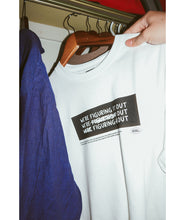 A photo of person hanging up a white t-shirt with a black rectangle in the center with the words "We're figuring it out" written three times in a row with the second line has "figuring" crossed out and the third line has "we're" and "it" crossed out. There is additional small text below this. From Answer in Progress.