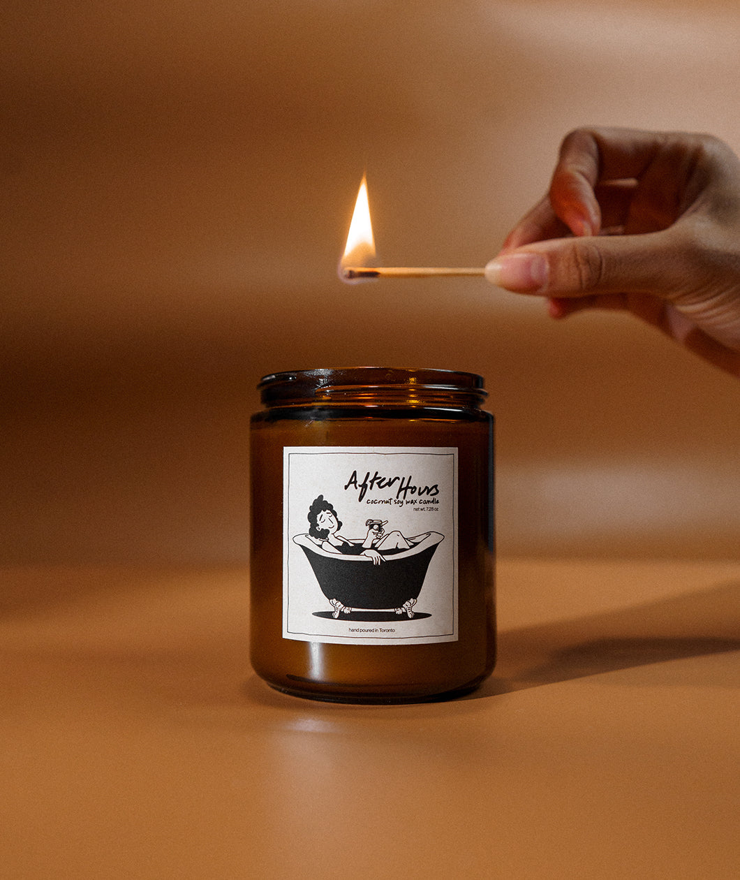 A candle about to be lit by a match. The candle’s label has a design of a person enjoying a bubble bath, with the title, “After Hours, coconut soy wax candle”. From Answer in Progress.