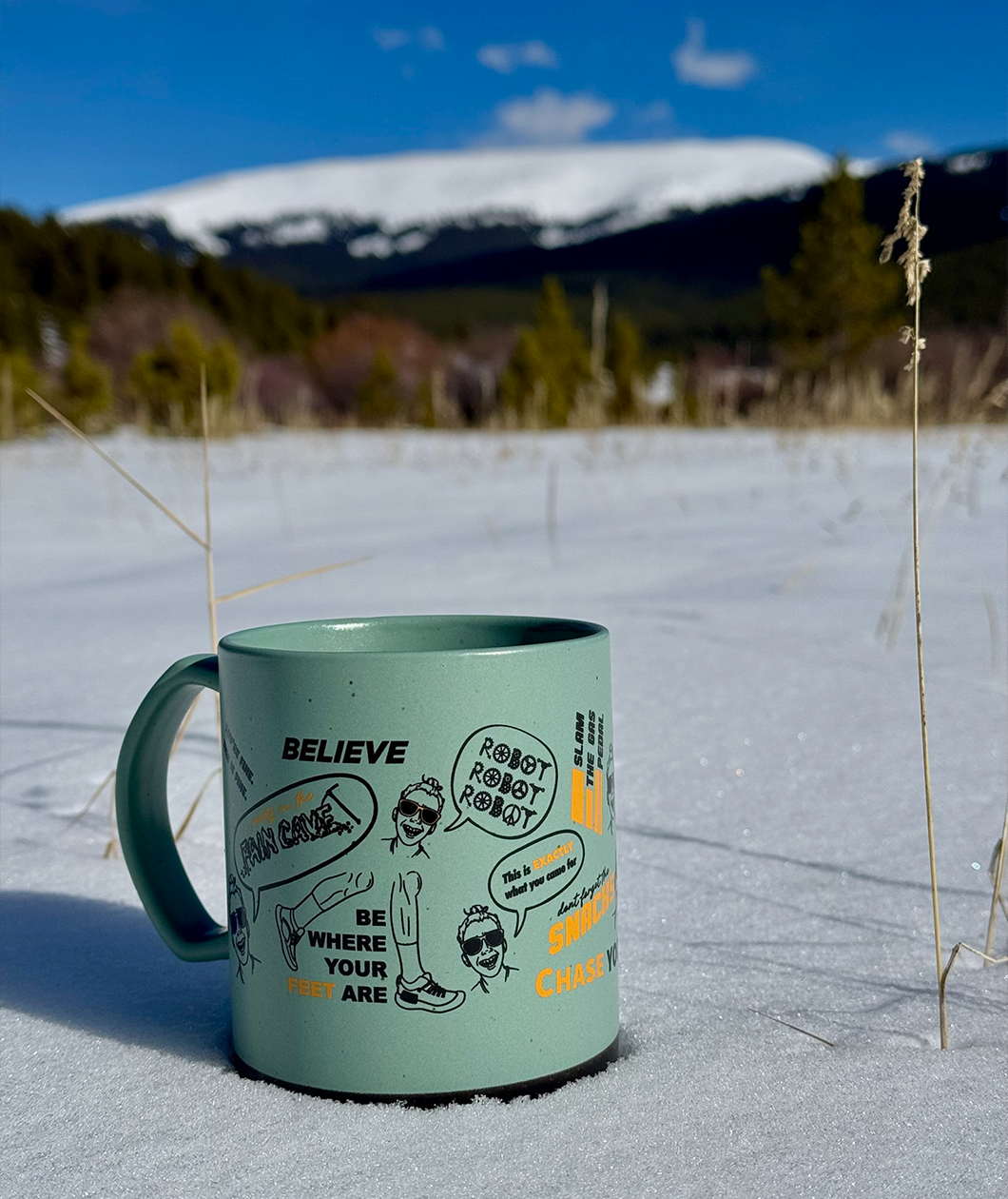 Green mug with black and orange printing of mantras around it sitting in the snow with a mountain behind it in the distance.