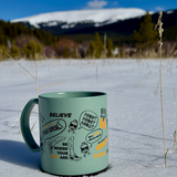 Green mug with black and orange printing of mantras around it sitting in the snow with a mountain behind it in the distance.