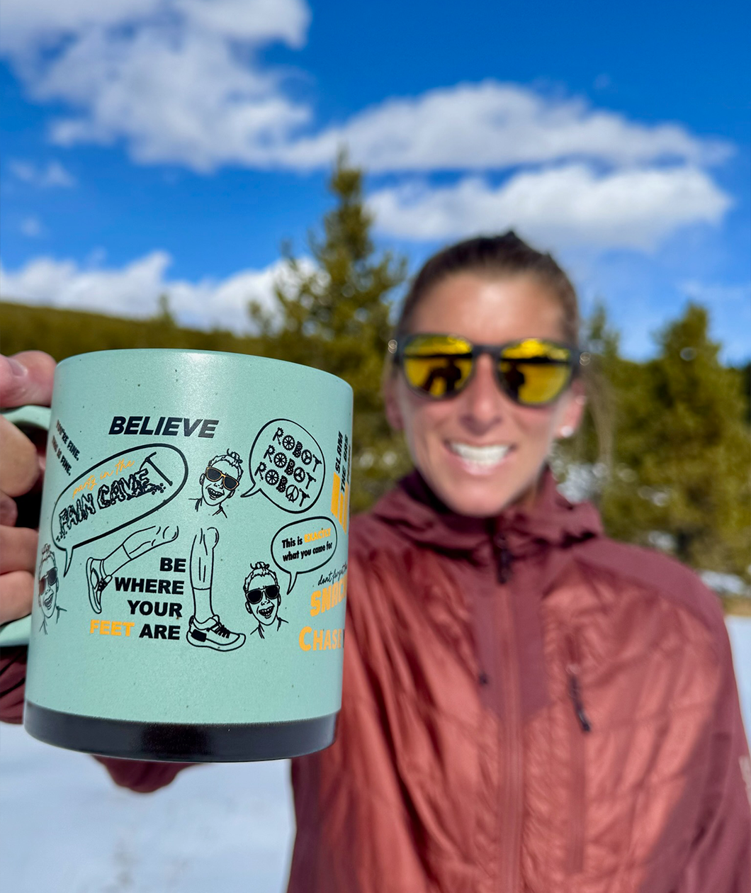 Courtney Dauwalter, an athletic woman wearing sunglasses and a red coat holding up a green mug to the the camera with black and orange printing around it with her mantras and illustrations of her.