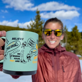 Courtney Dauwalter, an athletic woman wearing sunglasses and a red coat holding up a green mug to the the camera with black and orange printing around it with her mantras and illustrations of her.