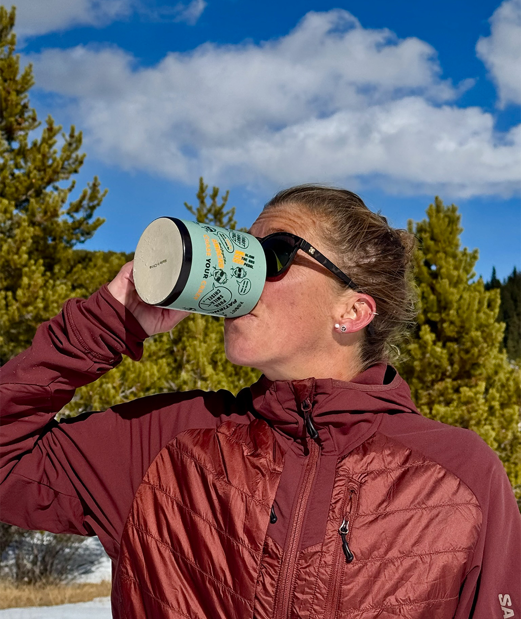 Courtney Dauwalter, athletic woman wearing sunglasses and a red coat drinking from a green mug with black and orange printing wrapped around it outside. The sky behind her is blue with some clouds and pine trees.