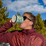 Courtney Dauwalter, athletic woman wearing sunglasses and a red coat drinking from a green mug with black and orange printing wrapped around it outside. The sky behind her is blue with some clouds and pine trees.