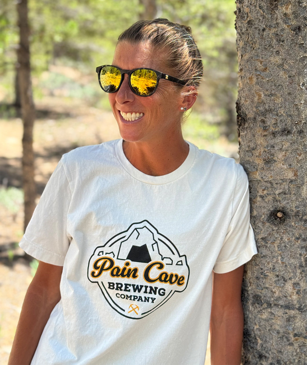 Courtney Dauwalter is leaning against a tree and smiling. She is wearing sunglasses and an off-white t-shirt with a design of a fake brewing company logo on the front. The logo is of a cave with the text, "Pain Cave Brewing Company" beneath it. From Courtney Dauwalter.