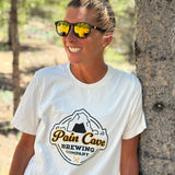 Courtney Dauwalter is leaning against a tree and smiling. She is wearing sunglasses and an off-white t-shirt with a design of a fake brewing company logo on the front. The logo is of a cave with the text, "Pain Cave Brewing Company" beneath it. From Courtney Dauwalter.
