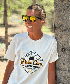 Courtney Dauwalter is leaning against a tree and smiling. She is wearing sunglasses and an off-white t-shirt with a design of a fake brewing company logo on the front. The logo is of a cave with the text, "Pain Cave Brewing Company" beneath it. From Courtney Dauwalter.