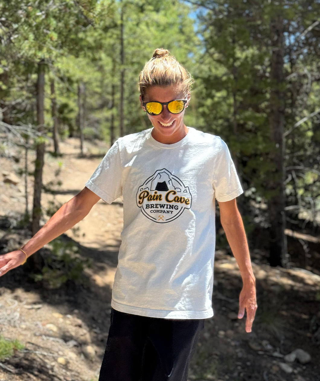 Courtney Dauwalter is walking through a forested trail and smiling. She is wearing sunglasses and an off-white t-shirt with a design of a fake brewing company logo on the front. The logo is of a cave with the text, "Pain Cave Brewing Company" beneath it. From Courtney Dauwalter.