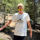 Courtney Dauwalter is walking through a forested trail and smiling. She is wearing sunglasses and an off-white t-shirt with a design of a fake brewing company logo on the front. The logo is of a cave with the text, "Pain Cave Brewing Company" beneath it. From Courtney Dauwalter.