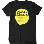 A mockup of a black t-shirt with a design featuring a yellow and black paper cut out Dear Hank & John logo. It has been modified to read "Dear John + Hank"