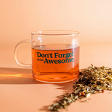 Clear glass mug with the words "*Don't Forget to be Awesome" on it in teal text. Sitting next to pile of loose leaf tea, mug is filled with orange tea.