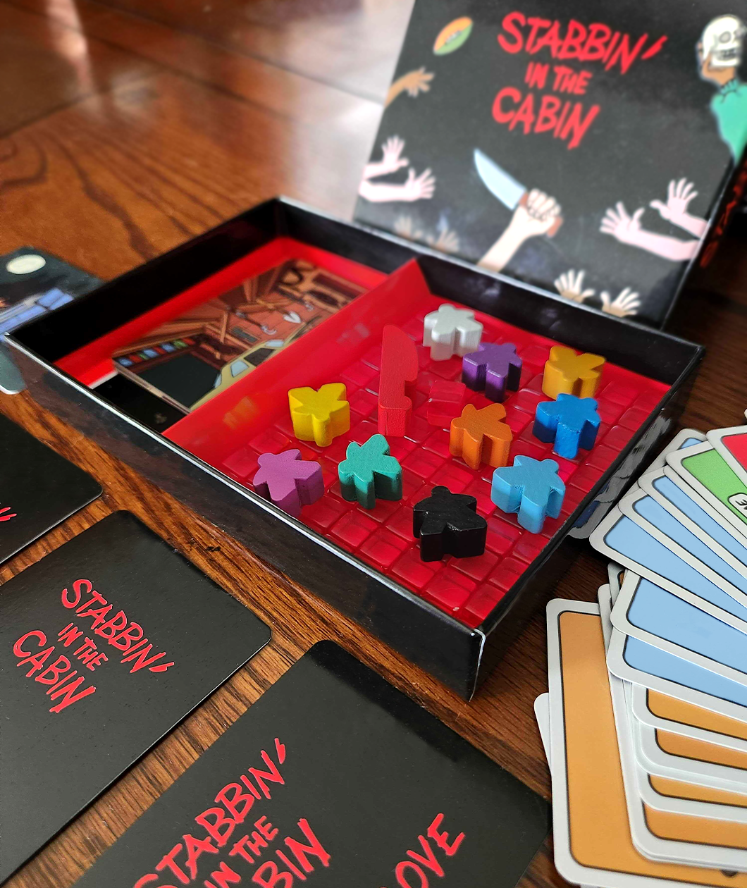 Open game box showing the bright red interior, red stab tokens, 10 colorful meeples, and the deck of cards with the front of the box in the background. Stabbin in the Cabin box is glossy black on the outside. There are some cards in the box and some cards fanned out around the box.