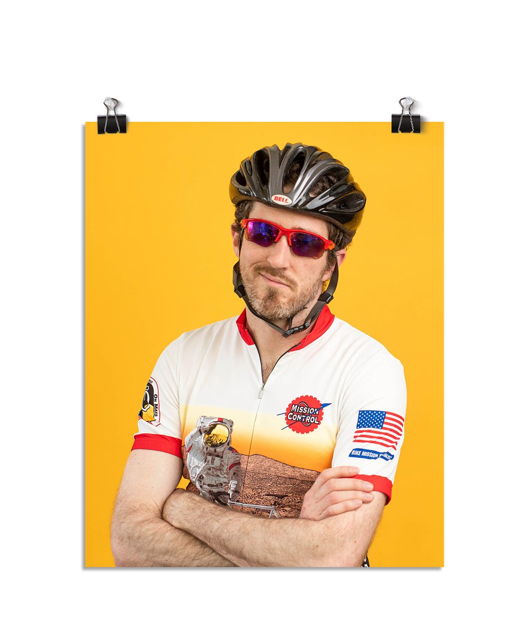 Photo of a man crossing his arms in a zip up bike shirt, bike helmet, and sun glasses smirking at the camera. On a yellow background.