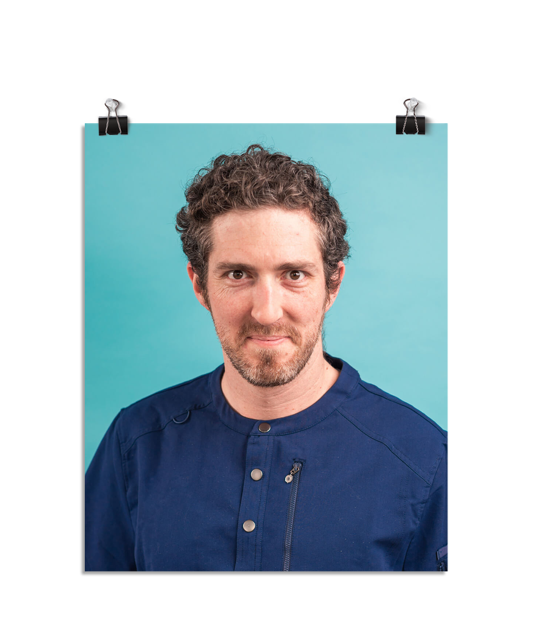 Photo of a man in navy blue scrubs with a soft smile and determined eyes that are a bit haunting. On a seafoam colored background.